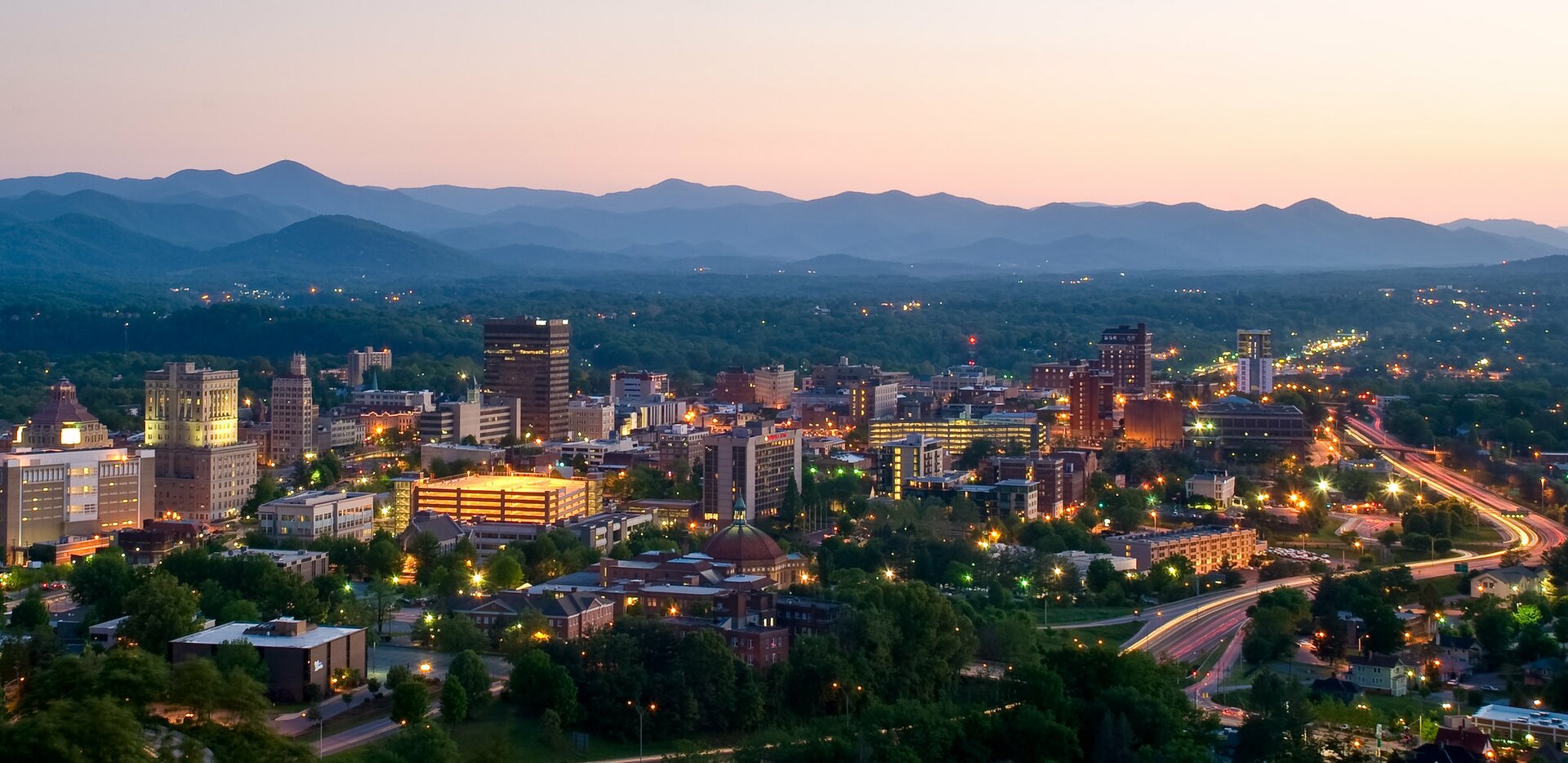 Aerial view of downtown Asheville, North Carolina at dusk with the Blue Ridge Mountains in the background